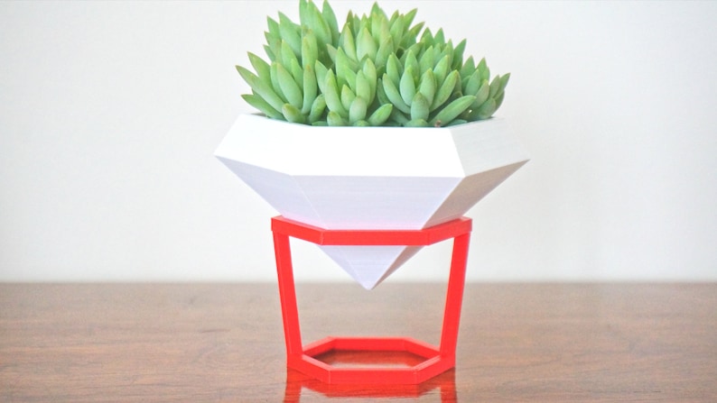 May include: A white geometric diamond-shaped planter with a succulent plant inside, sitting on a red metal stand.