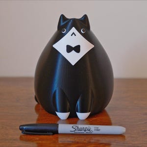 May include: A black cat figurine with a white bib and bow tie. The cat has white accents on its paws and eyes. A black Sharpie marker is in front of the cat on a wooden surface.