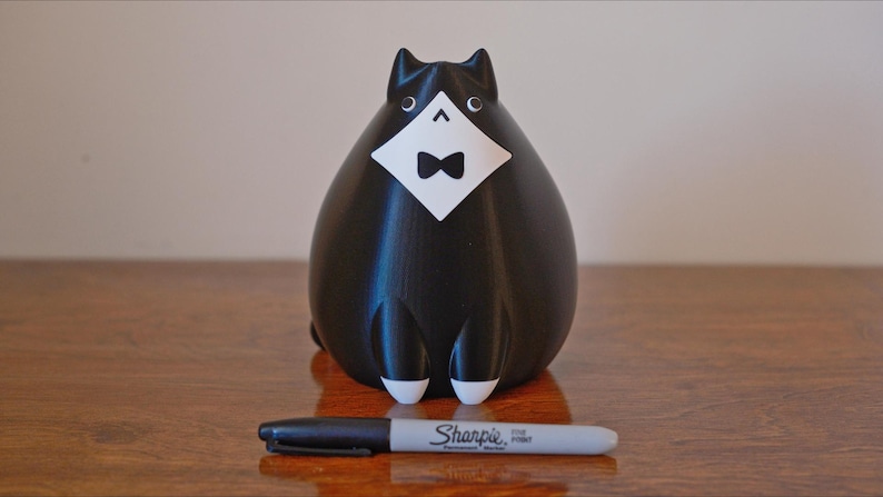 May include: A black cat figurine with a white bib and bow tie. The cat has white accents on its paws and eyes. A black Sharpie marker is in front of the cat on a wooden surface.