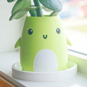 Puede incluir: Una maceta de cer&aacute;mica verde con forma de ping&uuml;ino de dibujos animados con la barriga blanca y una cara sonriente. La maceta est&aacute; sentada sobre un platillo blanco y tiene una peque&ntilde;a planta verde creciendo dentro.
