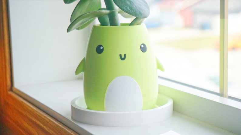 Puede incluir: Una maceta de cer&aacute;mica verde con forma de ping&uuml;ino de dibujos animados con la barriga blanca y una cara sonriente. La maceta est&aacute; sentada sobre un platillo blanco y tiene una peque&ntilde;a planta verde creciendo dentro.