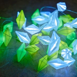 May include: A string of decorative lights featuring geometric shapes in shades of green and white. The lights are arranged on thin wires, creating a soft, ambient glow. The overall effect is a modern and whimsical aesthetic.