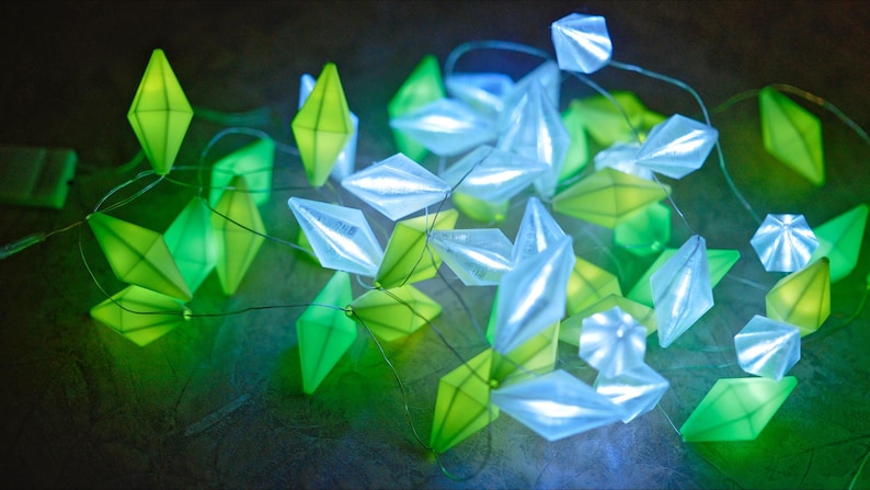 May include: A string of decorative lights featuring geometric shapes in shades of green and white. The lights are arranged on thin wires, creating a soft, ambient glow. The overall effect is a modern and whimsical aesthetic.