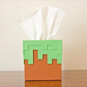 A cube tissue box cover inspired by the dirt grass block from the popular video game, Minecraft, is on a wooden table with a white background. It has bright green pixelated grass at the top half and the bottom half is brown.