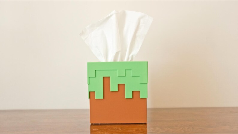 A cube tissue box cover inspired by the dirt grass block from the popular video game, Minecraft, is on a wooden table with a white background. It has bright green pixelated grass at the top half and the bottom half is brown.