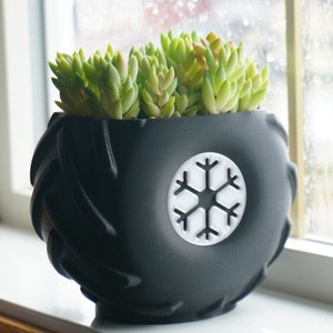May include: A black planter shaped like a tire with a white snowflake design. The planter is filled with green succulents.
