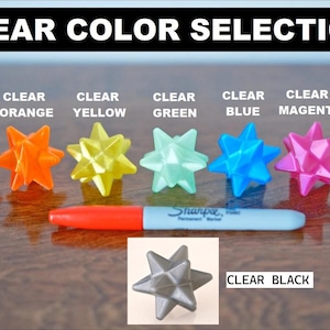 May include: Image showing a selection of clear, star-shaped ornaments in various colors: red, orange, yellow, green, blue, magenta, and white. A black Sharpie marker is in the center. The text "CLEAR COLOR SELECTION" is at the top.