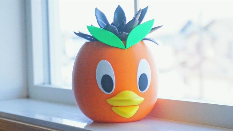 May include: Orange and yellow ceramic planter shaped like a cartoon duck with a succulent plant inside. The plant has dark green leaves and light green leaves.