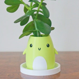 Puede incluir: Un macetero de dibujos animados verde y blanco con una cara sonriente y una peque&ntilde;a planta que crece en la parte superior. El macetero est&aacute; sentado sobre un platillo blanco.