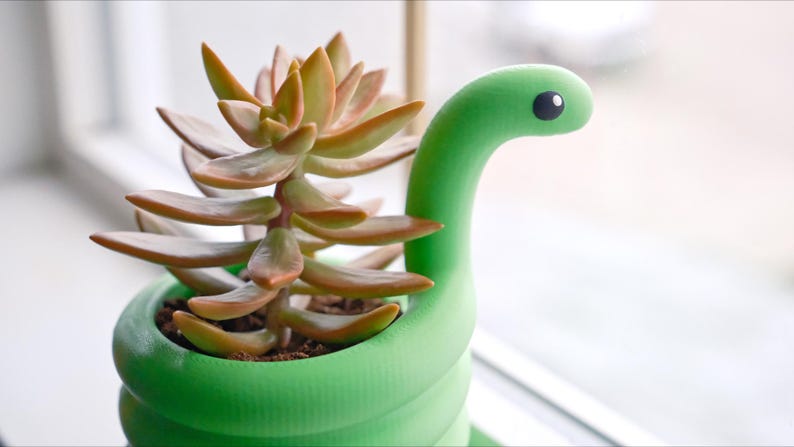 May include: A green snake-shaped planter holding a succulent plant. The succulent has fleshy, reddish-brown leaves. The planter has a whimsical design, with a snake head and a round eye. The planter is sitting on a windowsill.