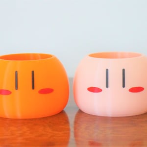 May include: Two ceramic dango-shaped planters, one orange and one pink, with black eyes and red cheeks.