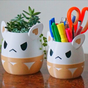 May include: Two white ceramic planters shaped like cartoon animals with angry expressions. The planter on the left has a small succulent plant inside. The planter on the right has a variety of colourful pens and a pair of orange scissors inside.