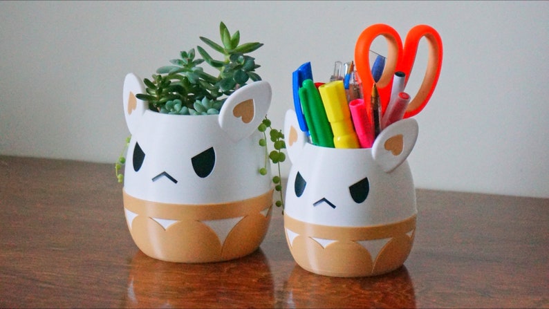 May include: Two white ceramic planters shaped like cartoon animals with angry expressions. The planter on the left has a small succulent plant inside. The planter on the right has a variety of colourful pens and a pair of orange scissors inside.
