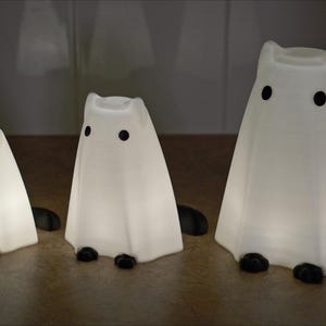 May include: Three white ghost-shaped night lights with black eyes. The ghosts are different sizes, with the largest ghost in the back and the smallest ghost in the front.