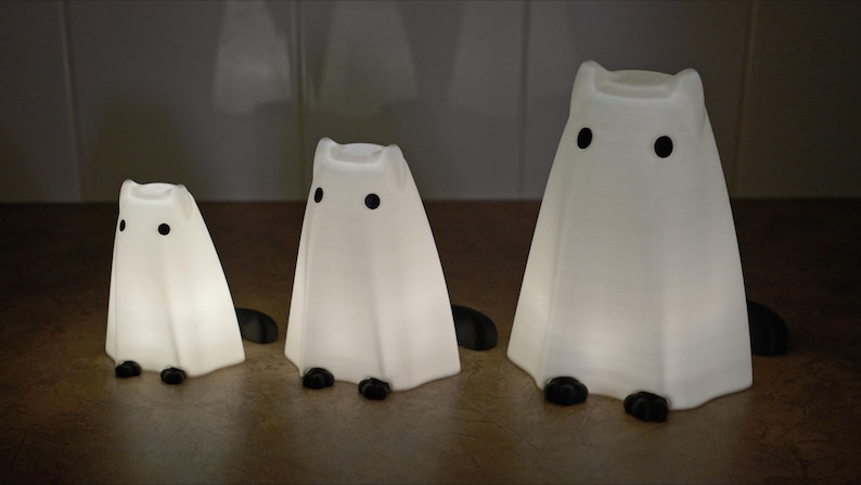 May include: Three white ghost-shaped night lights with black eyes. The ghosts are different sizes, with the largest ghost in the back and the smallest ghost in the front.