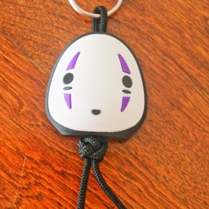 May include: A white and black keychain featuring a character face with purple accents. The keychain has a silver ring and a black cord. The design is a cartoon-style illustration.