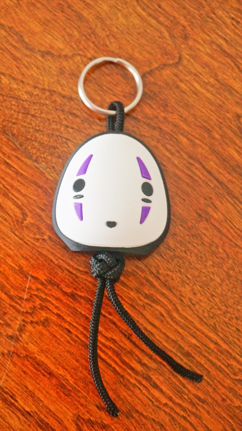 May include: A white and black keychain featuring a character face with purple accents. The keychain has a silver ring and a black cord. The design is a cartoon-style illustration.
