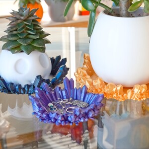 May include: Three white ceramic planters with different coloured crystal-like bases. The bases are blue, orange, and purple. The purple base has a few small pieces of jewellery inside.