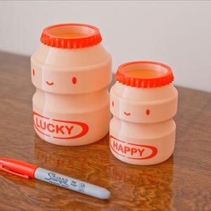 May include: Two light pink, cartoon-style bottles with orange lids, one labeled "LUCKY" and the other "HAPPY". A red and gray Sharpie marker lies in front of the bottles on a wooden surface. The bottles have smiling faces.