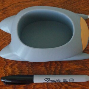 May include: A gray ceramic planter shaped like a cat with a light orange ear and a white paw. The planter has a round opening at the top for planting.