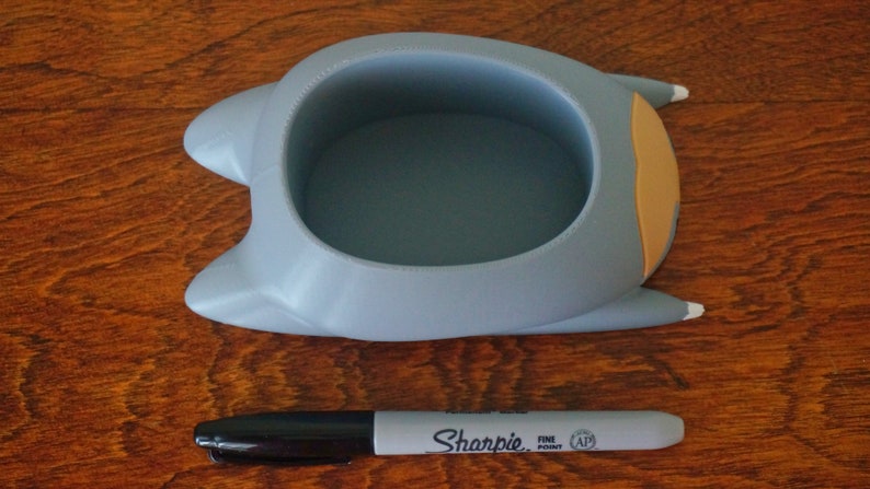 May include: A gray ceramic planter shaped like a cat with a light orange ear and a white paw. The planter has a round opening at the top for planting.