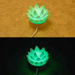 K&ouml;nnte beinhalten: Eine gr&uuml;ne, lotusbl&uuml;tenf&ouml;rmige Lampe. Die Lampe hat einen Holzsockel und ein wei&szlig;es Kabel. Die Bl&uuml;tenbl&auml;tter der Blume sind hellgr&uuml;n und die Lampe strahlt ein weiches Licht aus. Die Lampe ist f&uuml;r die Platzierung auf einer Oberfl&auml;che konzipiert.