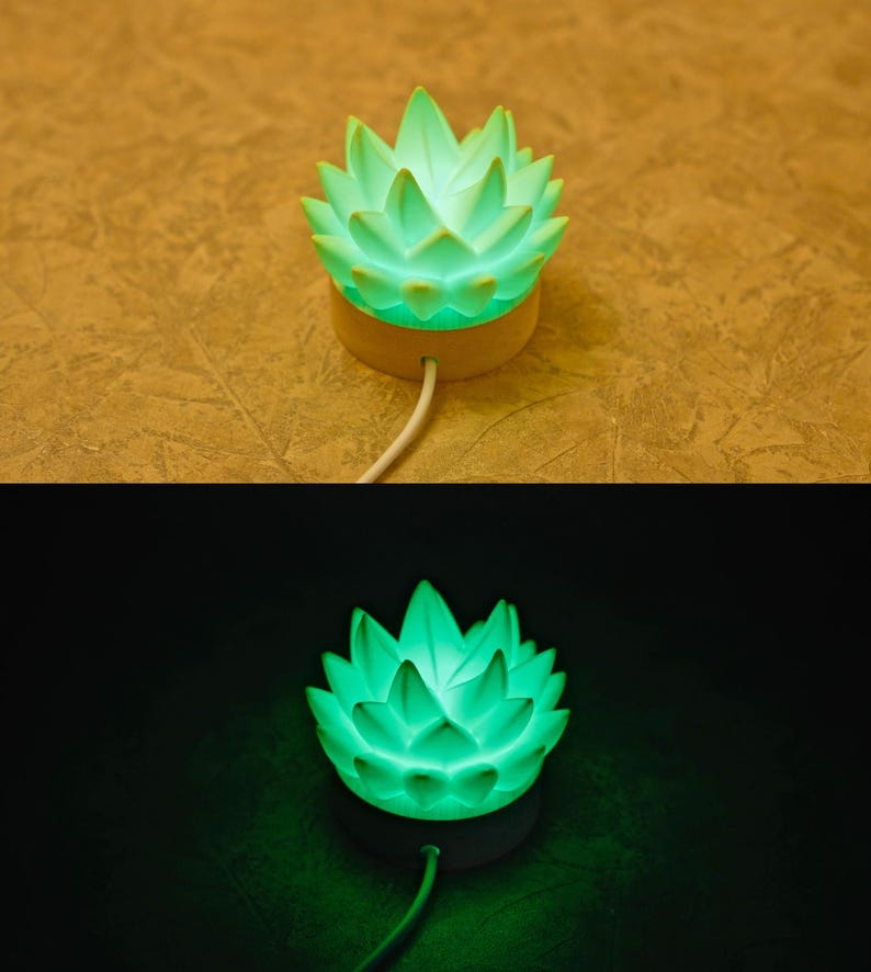 K&ouml;nnte beinhalten: Eine gr&uuml;ne, lotusbl&uuml;tenf&ouml;rmige Lampe. Die Lampe hat einen Holzsockel und ein wei&szlig;es Kabel. Die Bl&uuml;tenbl&auml;tter der Blume sind hellgr&uuml;n und die Lampe strahlt ein weiches Licht aus. Die Lampe ist f&uuml;r die Platzierung auf einer Oberfl&auml;che konzipiert.
