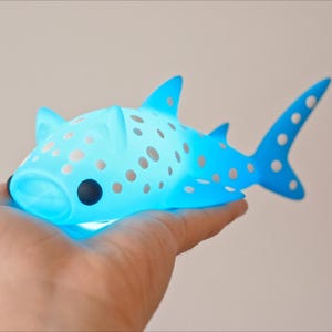 May include: A blue and white whale shark figurine with a light blue glow. The figurine is covered in white spots and has a cartoon-like face.