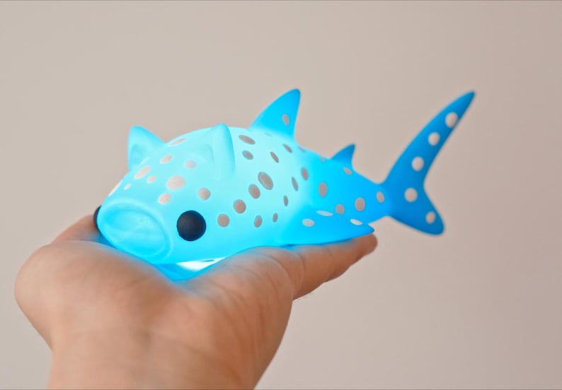May include: A blue and white whale shark figurine with a light blue glow. The figurine is covered in white spots and has a cartoon-like face.