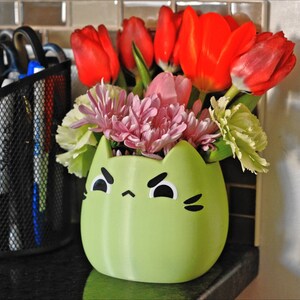 May include: A green ceramic cat-shaped vase with a grumpy face holds a bouquet of pink and white flowers, including tulips and carnations.