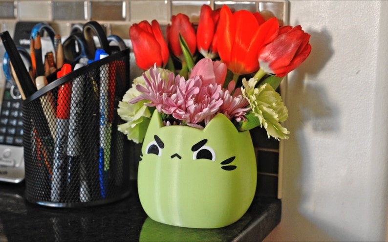 May include: A green ceramic cat-shaped vase with a grumpy face holds a bouquet of pink and white flowers, including tulips and carnations.