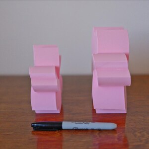 May include: Two pink paper sculptures, one larger than the other, stand on a wooden surface. The sculptures are made of folded paper and have a geometric shape.