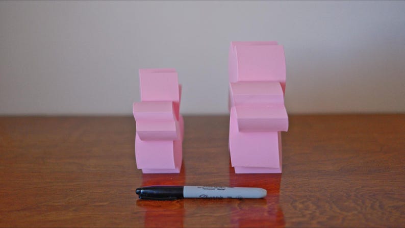 May include: Two pink paper sculptures, one larger than the other, stand on a wooden surface. The sculptures are made of folded paper and have a geometric shape.