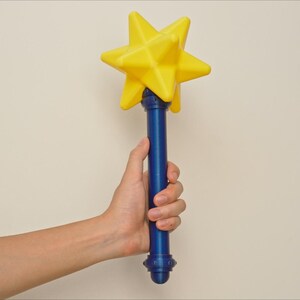 May include: A yellow star-shaped wand with a blue handle. The wand is held by a person. The star is a bright yellow color, and the handle is a deep blue. The wand has a decorative base.