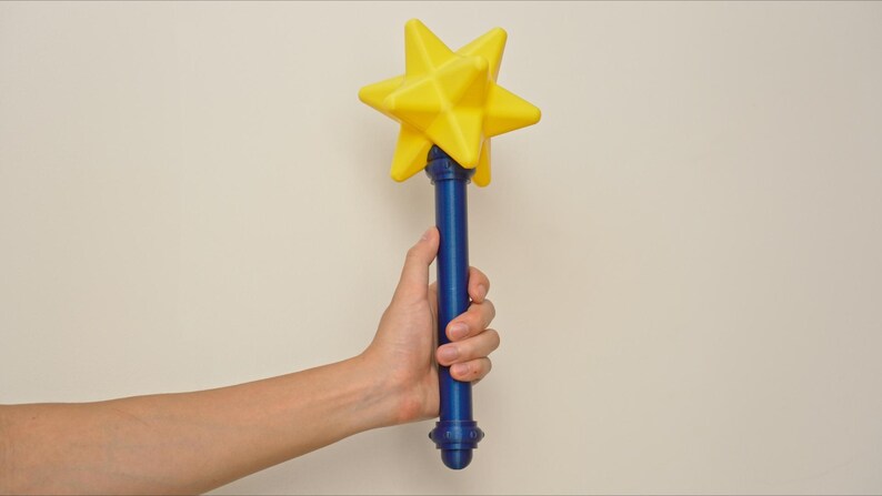 May include: A yellow star-shaped wand with a blue handle. The wand is held by a person. The star is a bright yellow color, and the handle is a deep blue. The wand has a decorative base.