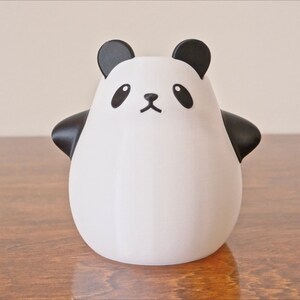 Op de afbeelding: Een wit keramiek panda-beeldje met zwarte oren, ogen, neus en mond. De panda staat op een houten oppervlak.