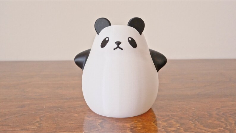 Op de afbeelding: Een wit keramiek panda-beeldje met zwarte oren, ogen, neus en mond. De panda staat op een houten oppervlak.