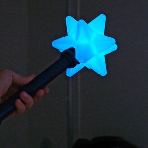 May include: A dark blue star-shaped wand with a glowing blue light. The wand is held in a hand against a dark background. The star has multiple points and a textured surface.