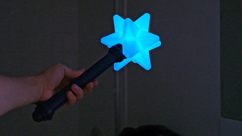 May include: A dark blue star-shaped wand with a glowing blue light. The wand is held in a hand against a dark background. The star has multiple points and a textured surface.