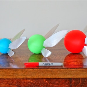 May include: Four different sized, round, hollow, plastic spheres with white wings attached to each. The spheres are pink, blue, green, and red. A white marker is in front of the spheres.