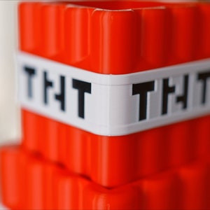 May include: A red plastic toy block with a white band that says "TNT" on it. The block is shaped like a Minecraft TNT block.