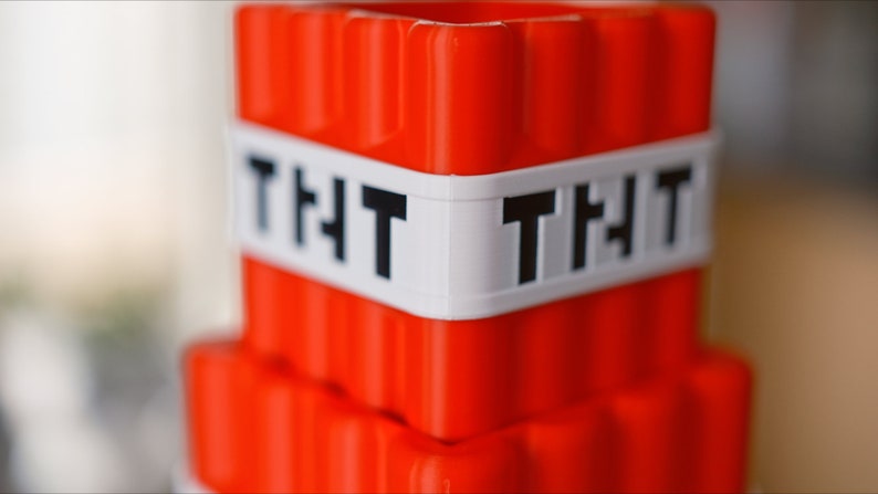 May include: A red plastic toy block with a white band that says "TNT" on it. The block is shaped like a Minecraft TNT block.