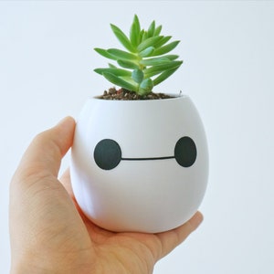 Pu&ograve; includere: Un vaso di ceramica bianco a forma di robot dei cartoni animati con occhi neri e bocca, che contiene una piccola pianta grassa verde.