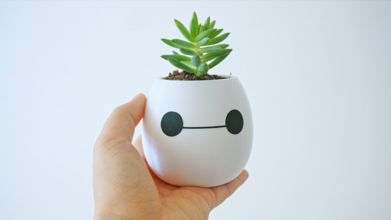 Pu&ograve; includere: Un vaso di ceramica bianco a forma di robot dei cartoni animati con occhi neri e bocca, che contiene una piccola pianta grassa verde.