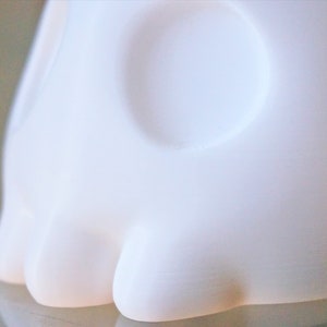 May include: White ceramic sculpture of a skull with a smooth, rounded surface. The skull has two large eye sockets and a prominent nose.
