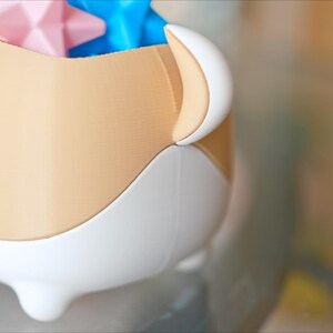 May include: A beige and white ceramic container with a rounded shape and three legs. The container is filled with colorful star-shaped toys.