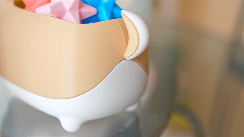 May include: A beige and white ceramic container with a rounded shape and three legs. The container is filled with colorful star-shaped toys.