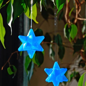 May include: Two blue, star-shaped ornaments hang from a leafy green plant. The stars are made of a translucent material and have a 3D geometric design.