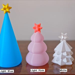 May include: Three decorative Christmas trees in light blue, light pink, and white. The light blue tree is cone-shaped with a yellow star. The light pink tree is rounded with an orange star. The white tree is geometric with a white star.