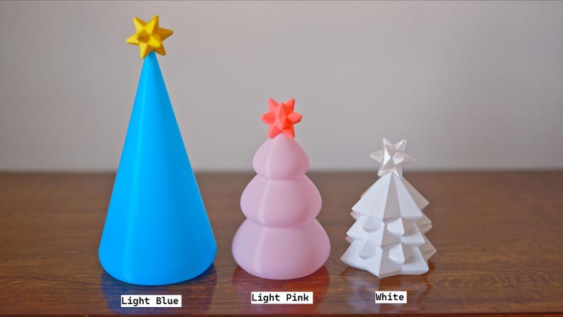 May include: Three decorative Christmas trees in light blue, light pink, and white. The light blue tree is cone-shaped with a yellow star. The light pink tree is rounded with an orange star. The white tree is geometric with a white star.